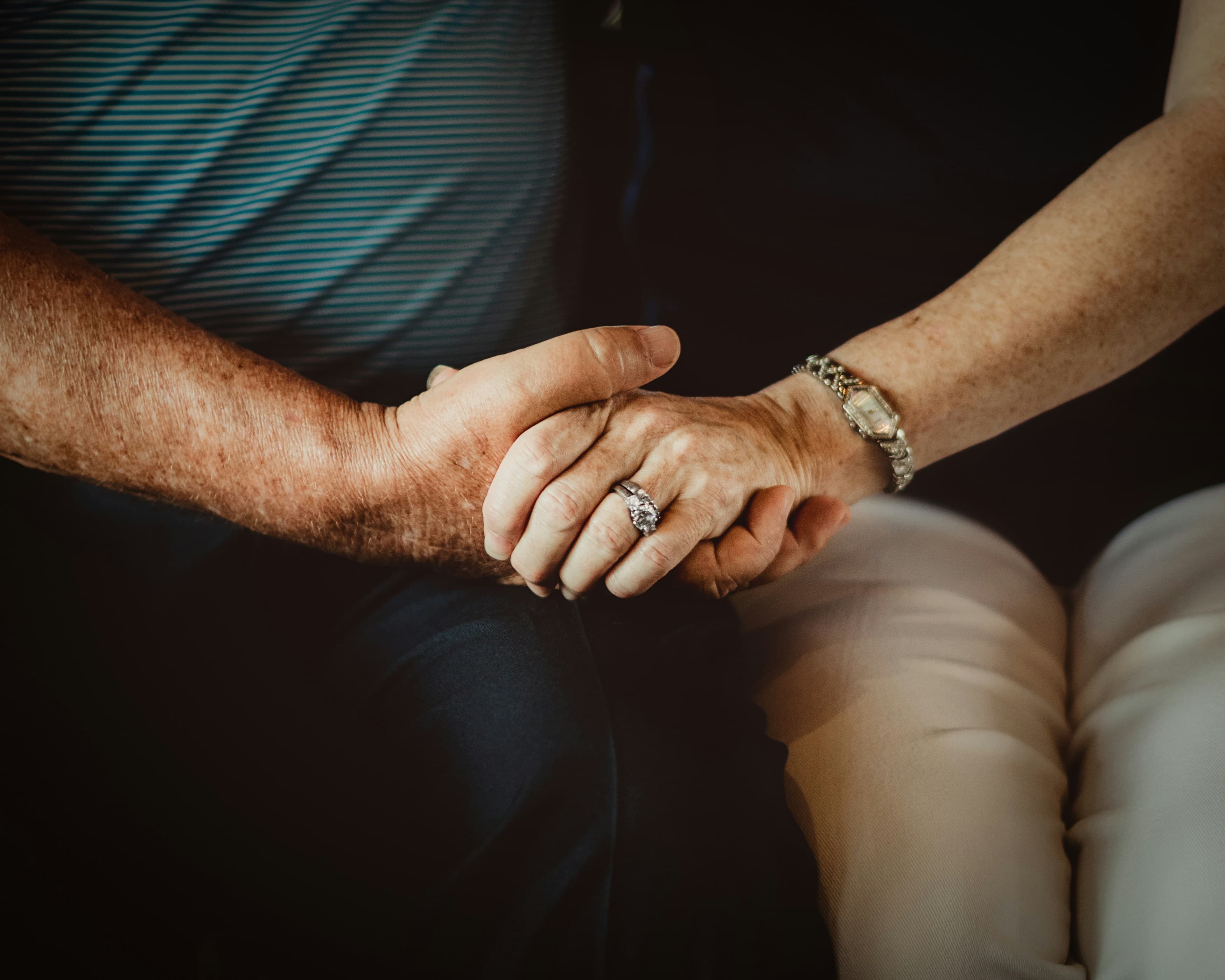 An older couple's hands clasped together, wedding band visible, the weight of a long marriage