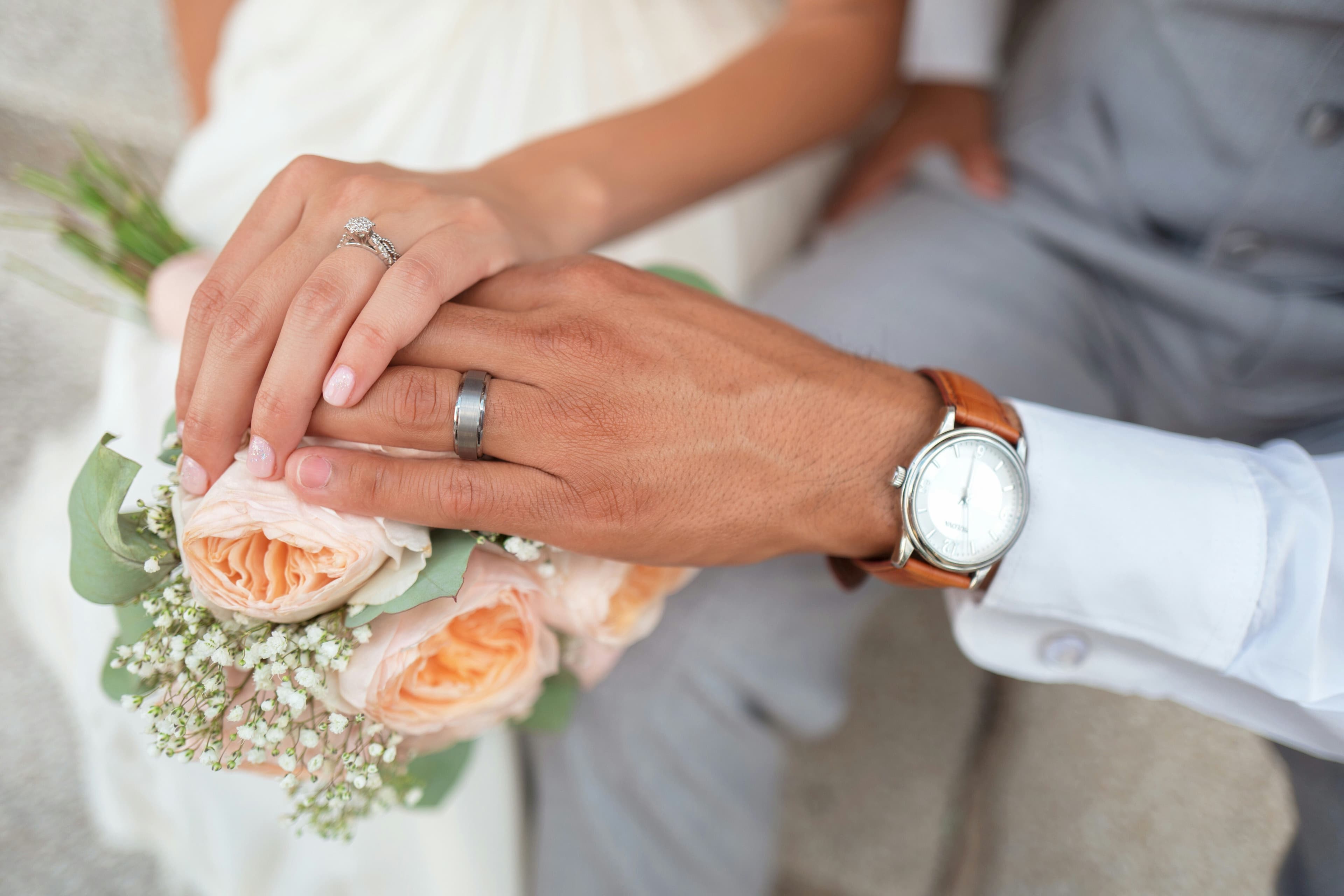 Bride and groom's hands resting on a peach-rose bouquet, both wearing rings